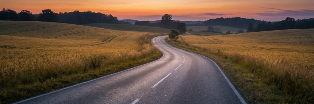 Carretera rural vacía al atardecer con curvas entre campos, ideal para ilustrar viajes y preparación ante imprevistos.